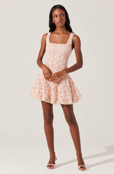A woman is wearing a sleeveless, light pink dress with a textured floral lace overlay. The dress has wide shoulder straps, a fitted bodice, and a flared skirt that falls above the knees. She is accessorized with gold earrings, a gold bracelet, and a gold ring, and is wearing matching light pink open-toe heels.